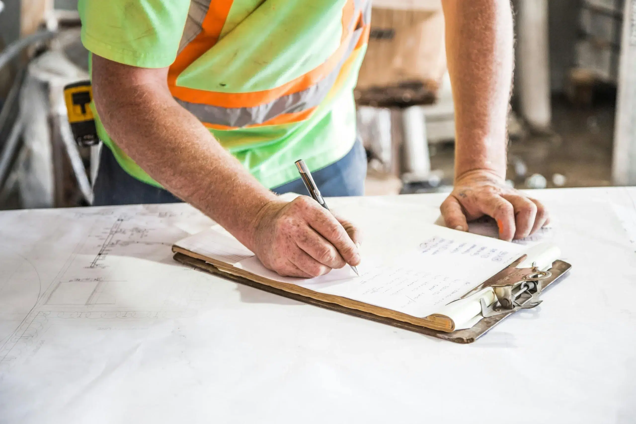 A maintenance technician in a high-visibility vest taking notes on a clipboard over technical blueprints.