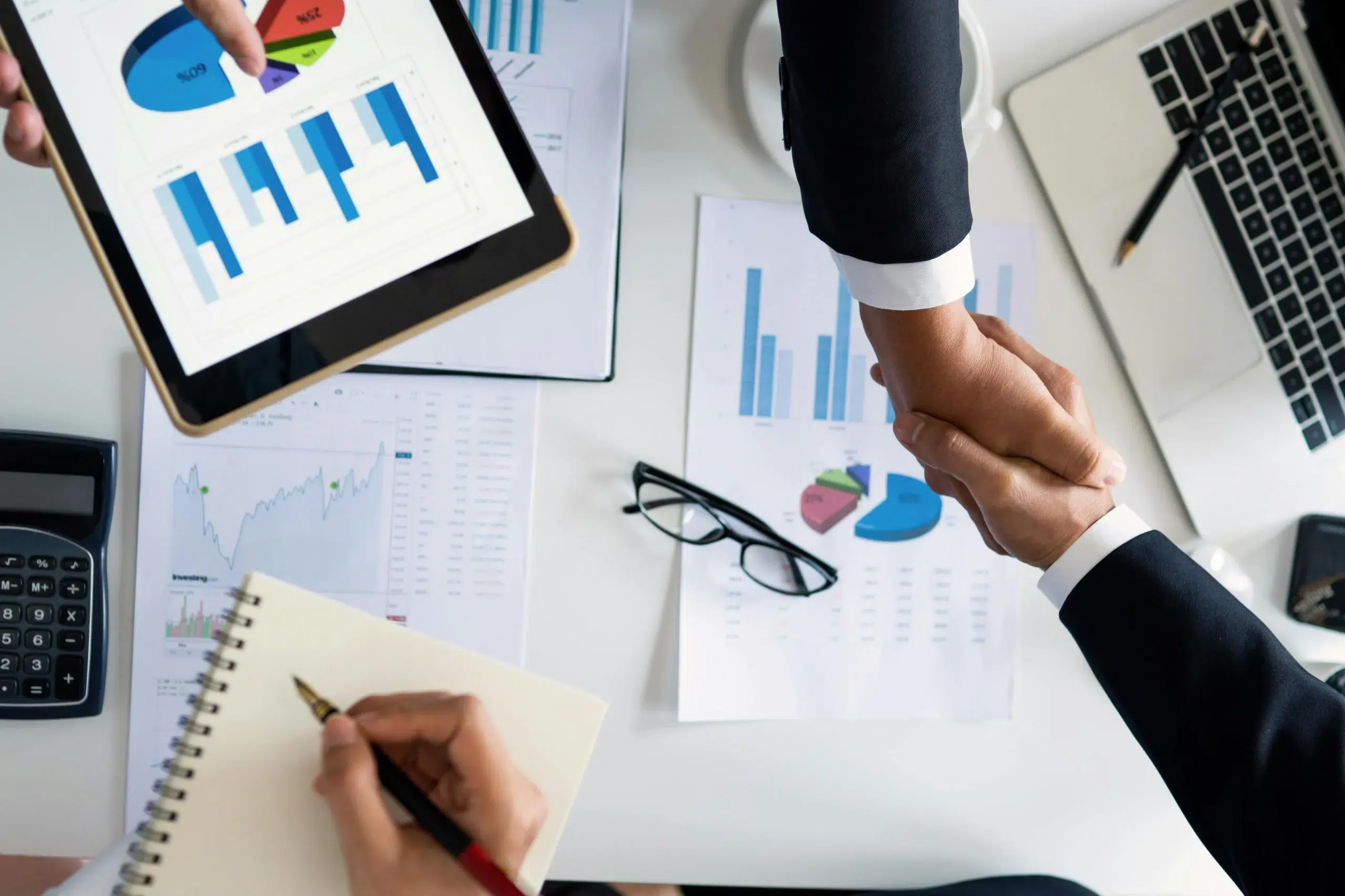 Two business professionals shaking hands over a desk with financial charts, symbolizing the integration between ERP financial visibility and CMMS operational depth.