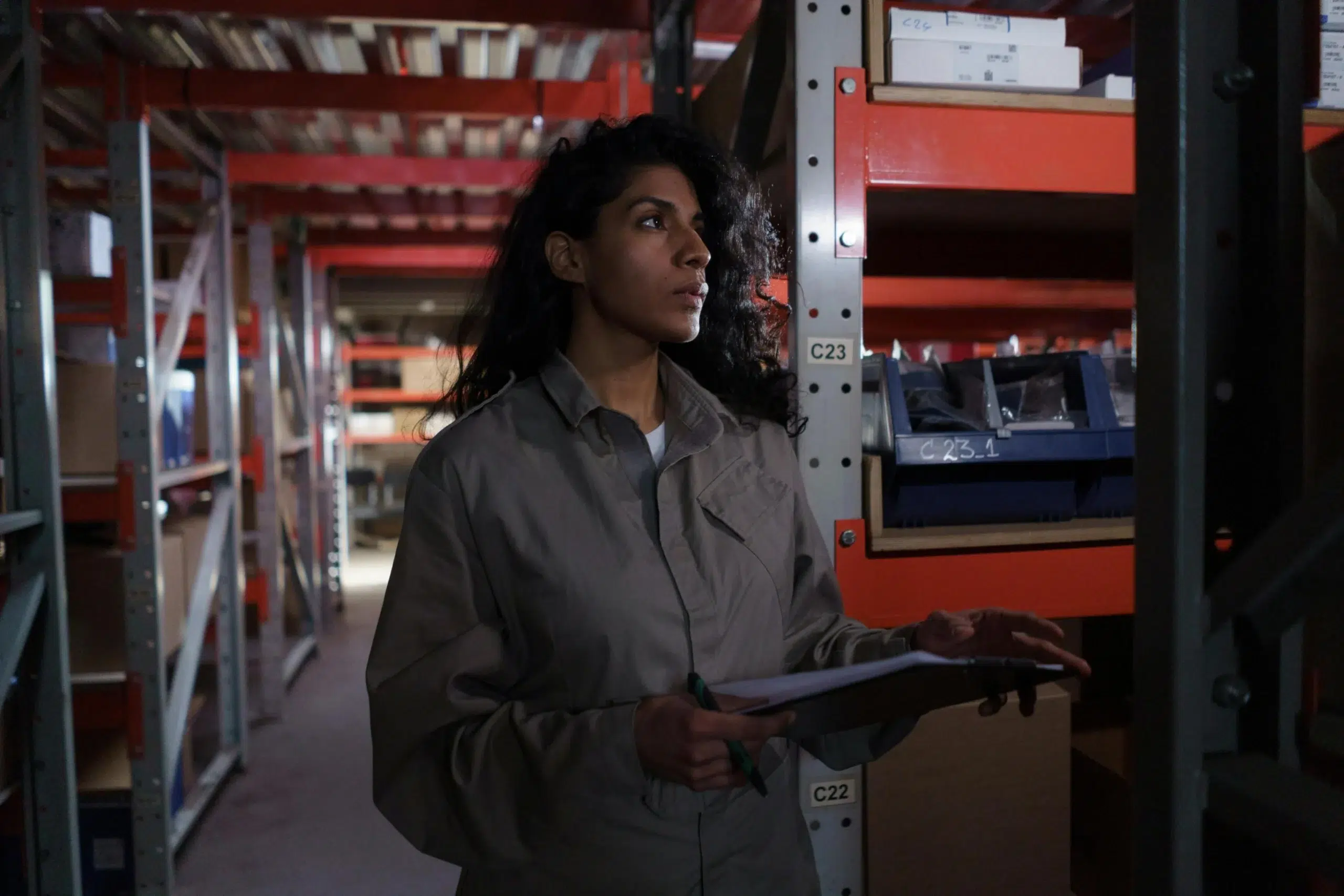 A female maintenance worker in a jumpsuit holding a clipboard and inspecting stock levels in a dimly lit industrial warehouse.