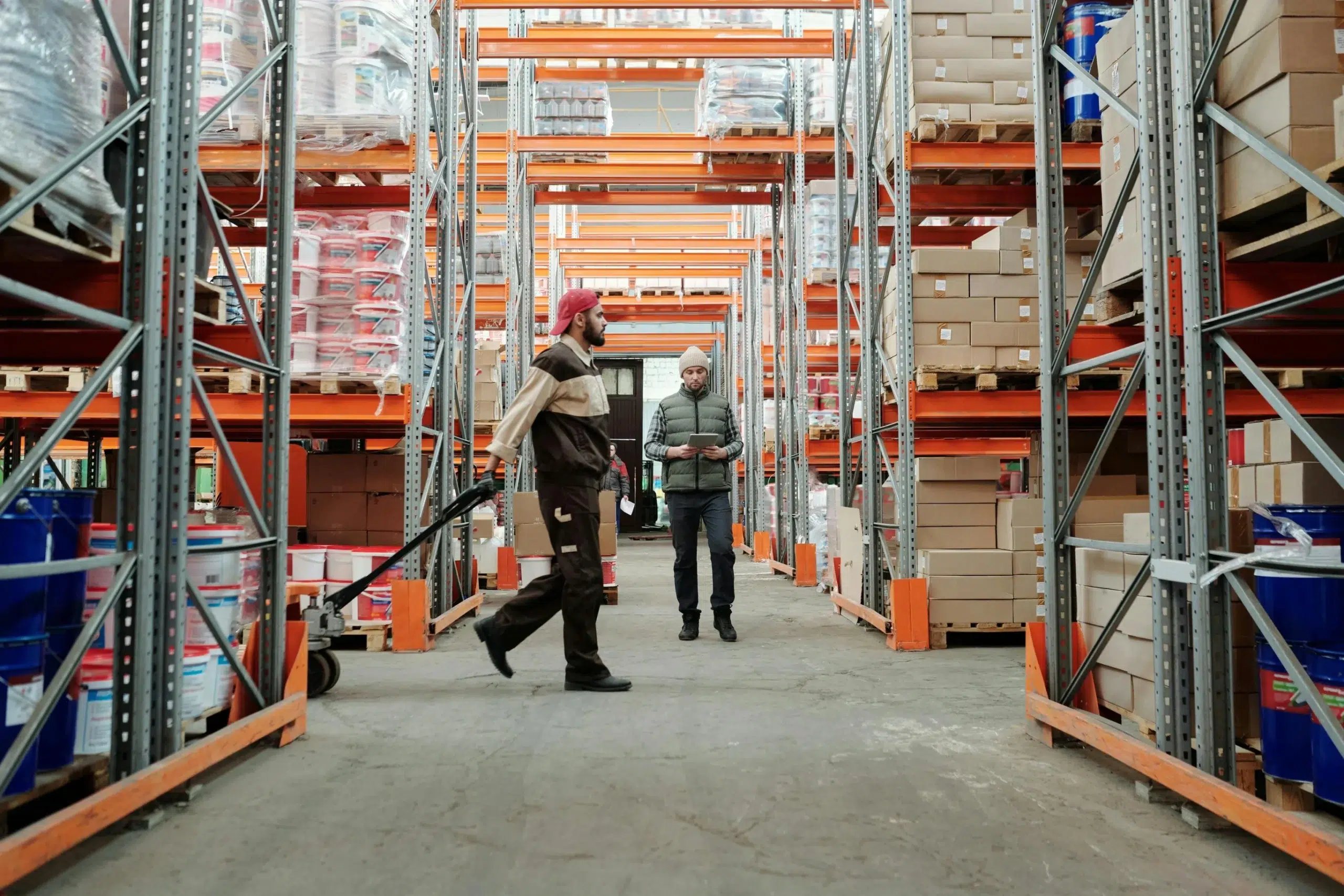 Warehouse workers managing stock in an industrial facility with high-density shelving and pallet jacks.