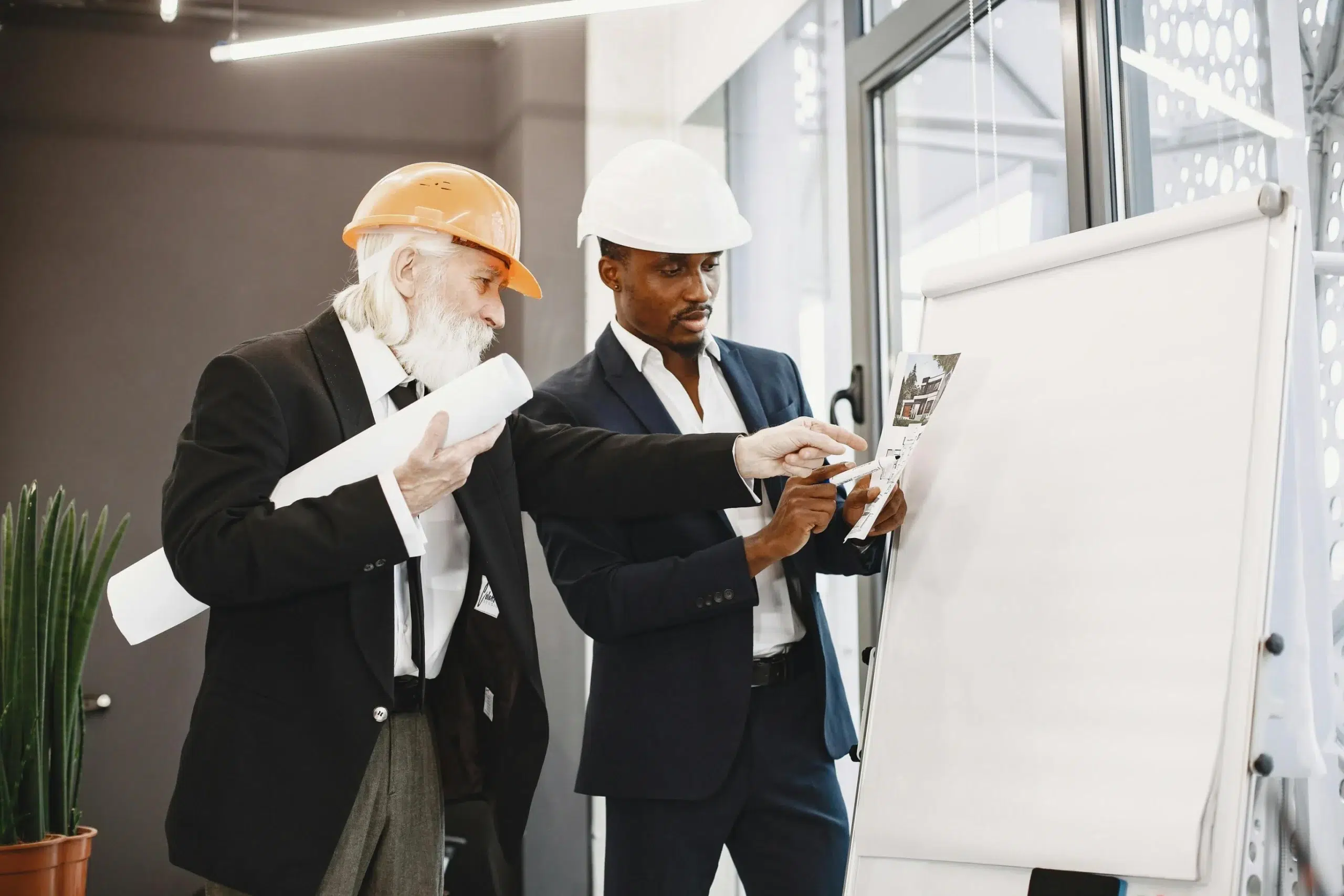 Two professionals in hard hats, one an older executive and one a younger manager, reviewing architectural plans and KPIs on a whiteboard.